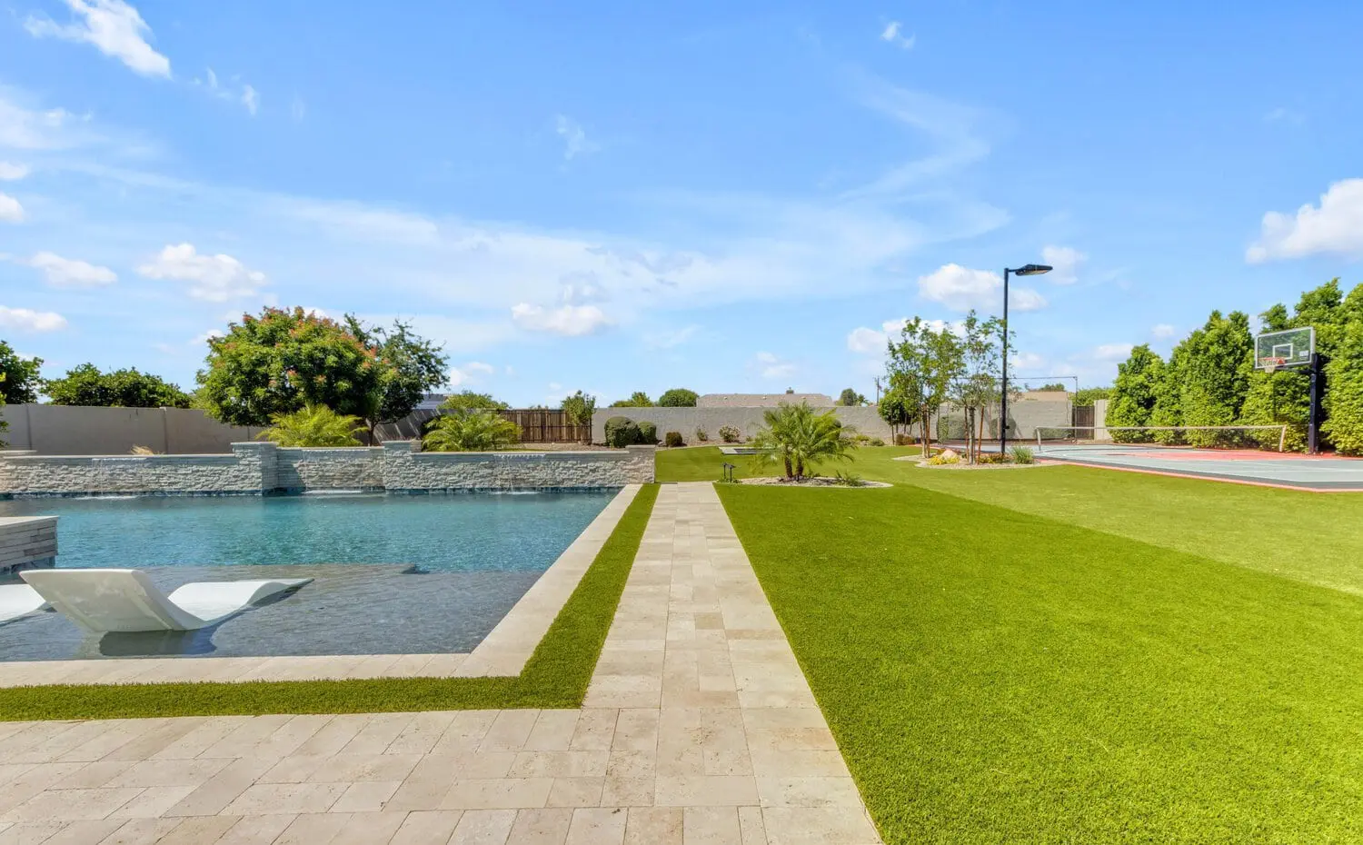 A serene Tucson, AZ backyard scene features a swimming pool with in-water chaise lounges, adjacent to a manicured lawn of synthetic turf. A basketball hoop is visible on a court in the background under the clear blue sky.