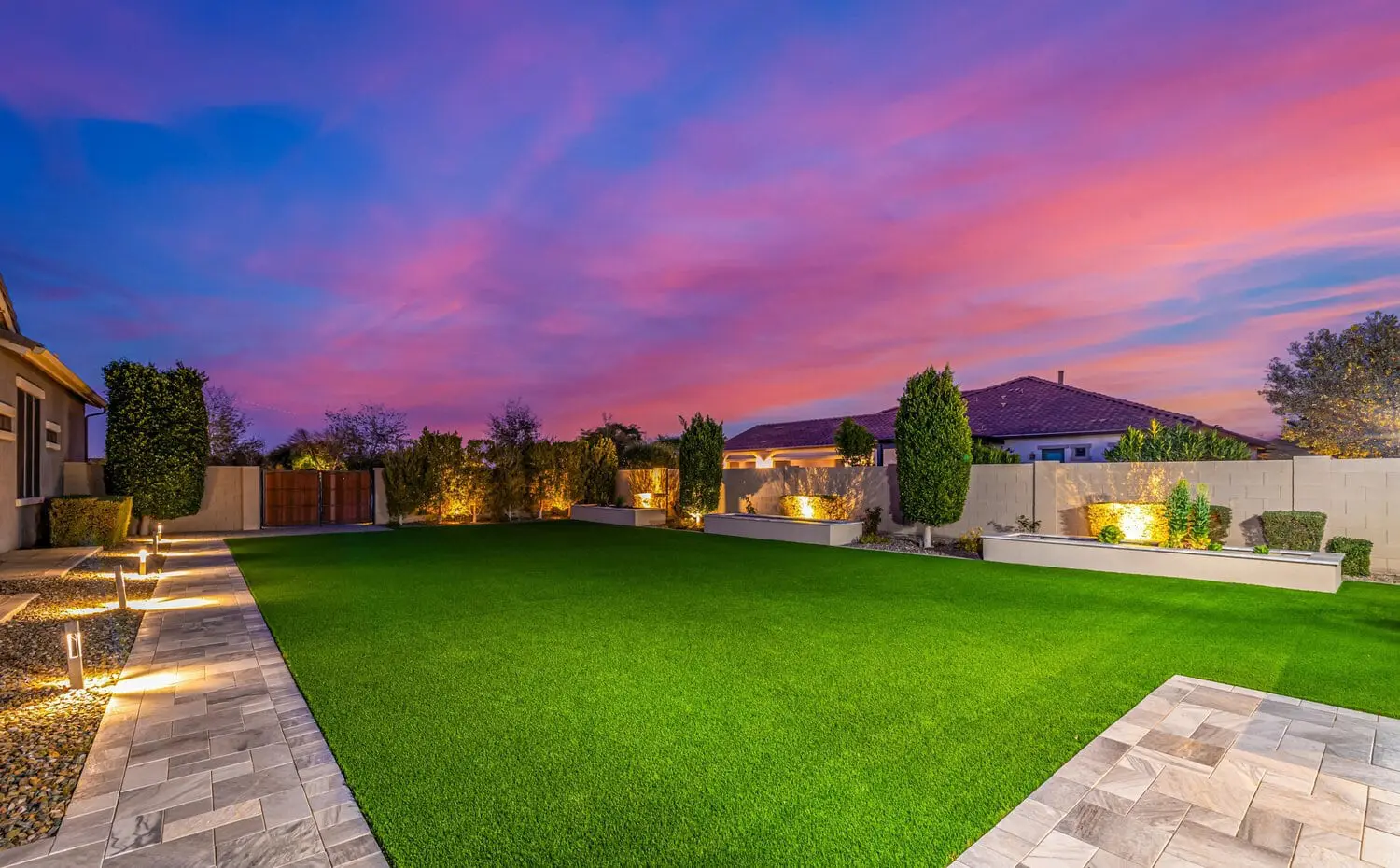 A backyard at sunset in Tucson, AZ boasts a vibrant pink and purple sky. The yard features synthetic turf, bordered by tiled pathways, small trees, and hedges. Soft lighting illuminates the garden beds and pathways.