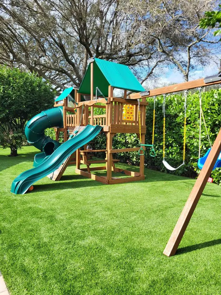 A backyard playground featuring a wooden playset with a green slide, covered platforms, and swings. The playset is surrounded by lush Scottsdale Turf Pros’ artificial grass installation, with tall trees in the background, creating a vibrant and inviting outdoor play area for children in Phoenix, AZ.