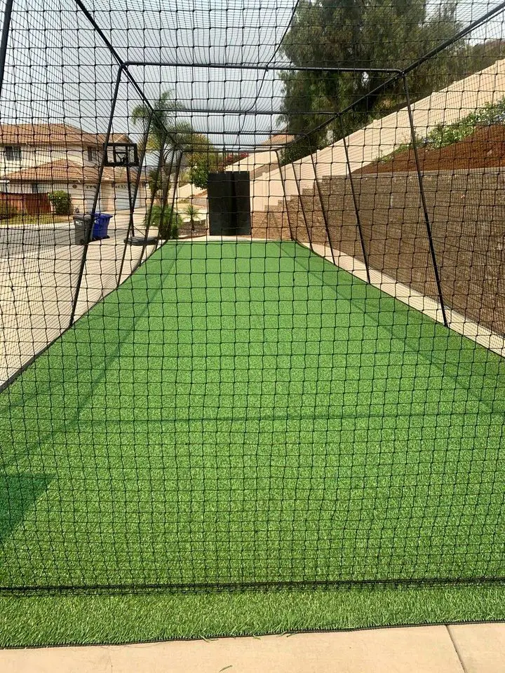 A rectangular outdoor sports net enclosure features artificial green turf, ensuring a maintenance-free lawn. The enclosure, with black netting and a metallic frame, extends along a pathway surrounded by a beige wall and residential houses. Trees and a basketball hoop are visible in the background.