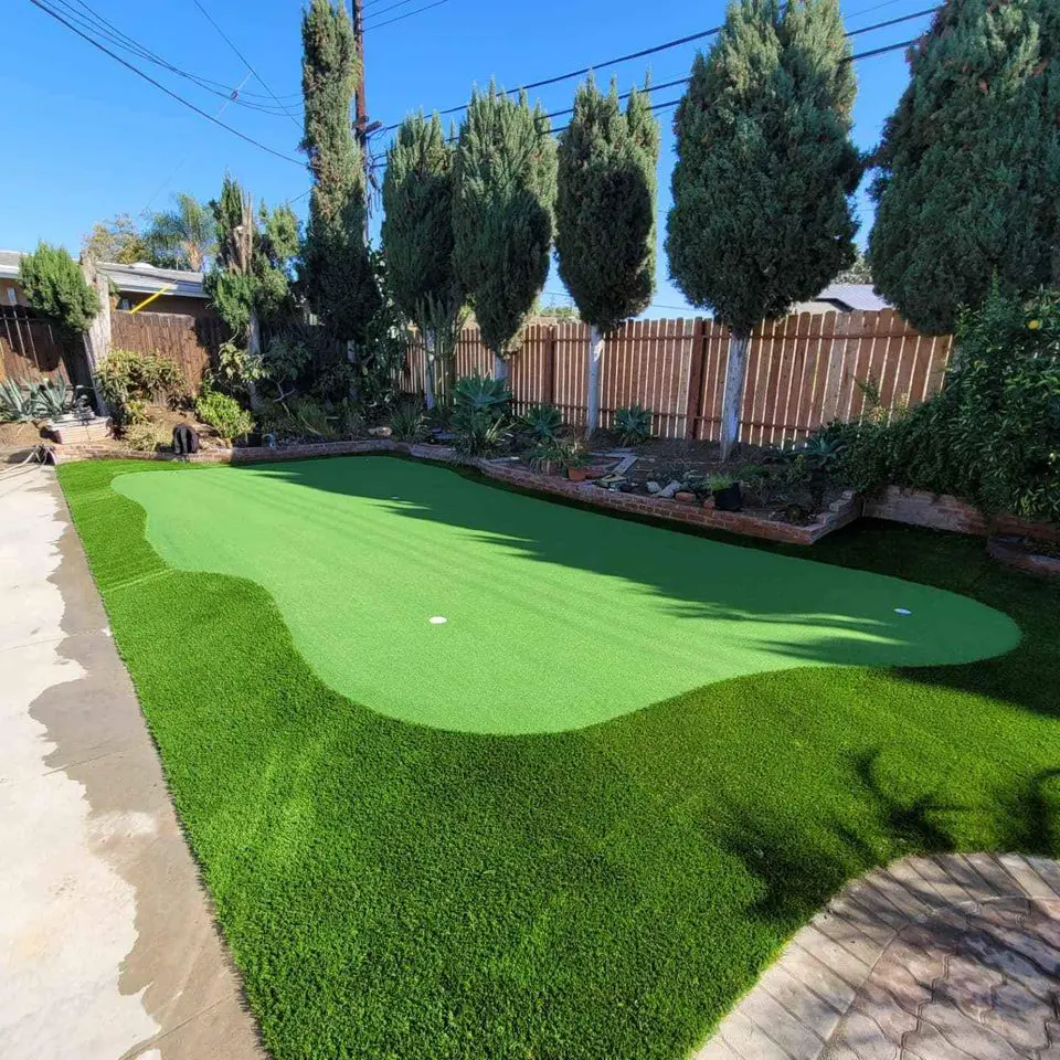 A backyard oasis features backyard golf greens with two holes, enveloped by vibrant artificial turf. Tall, lush trees line the border against a wooden fence under a clear blue sky.