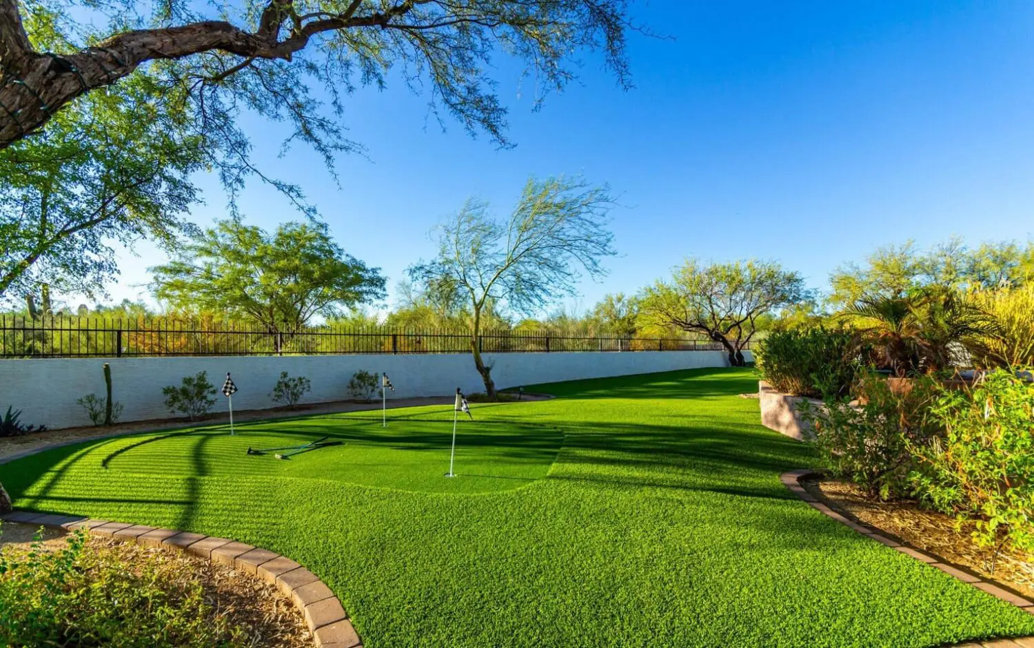 A lush backyard in Tucson, AZ, features a well-maintained mini golf course with synthetic grass solutions. Surrounded by trees, a stone path, and a white fence under a clear blue sky, it creates a serene outdoor atmosphere enhanced by cutting-edge artificial turf installations.