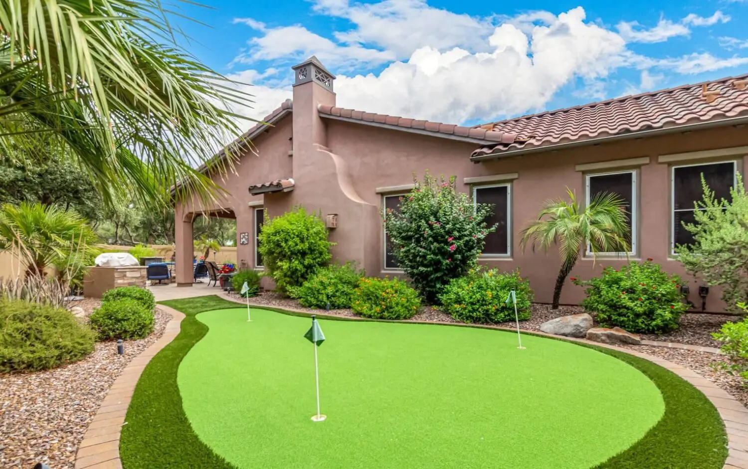 A backyard golf paradise features a lush putting green nestled among desert landscaping, with palm trees and shrubs. The terracotta-colored house with a tiled roof stands proudly in the background, all beneath a partly cloudy sky.