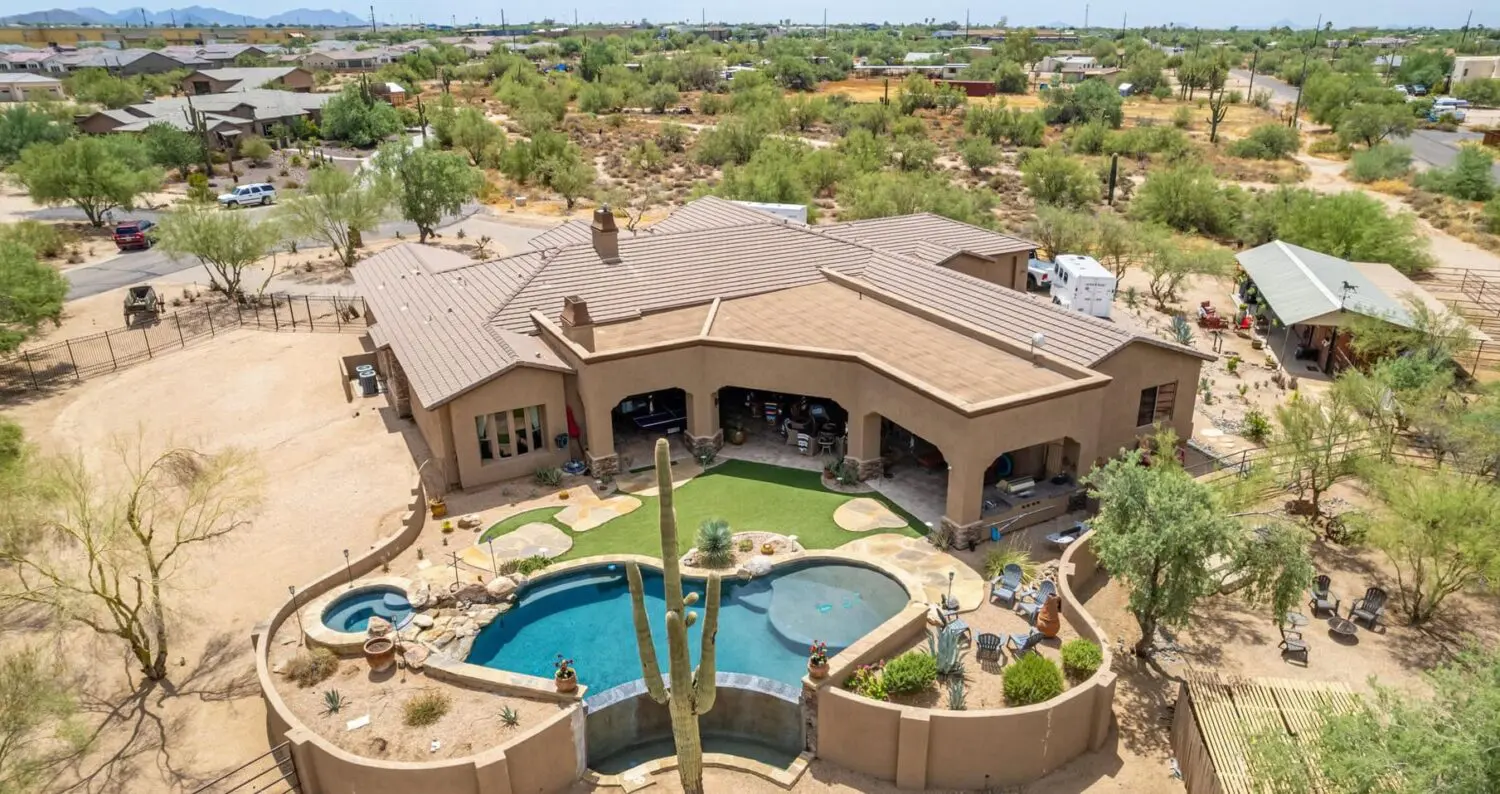 Aerial view of a large, southwestern-style house in Tucson, AZ, with a beige stucco exterior. The backyard boasts a pool with a cactus sculpture, lounging areas, and synthetic turf. Set in a desert landscape with scattered bushes and neighboring houses in the distance.