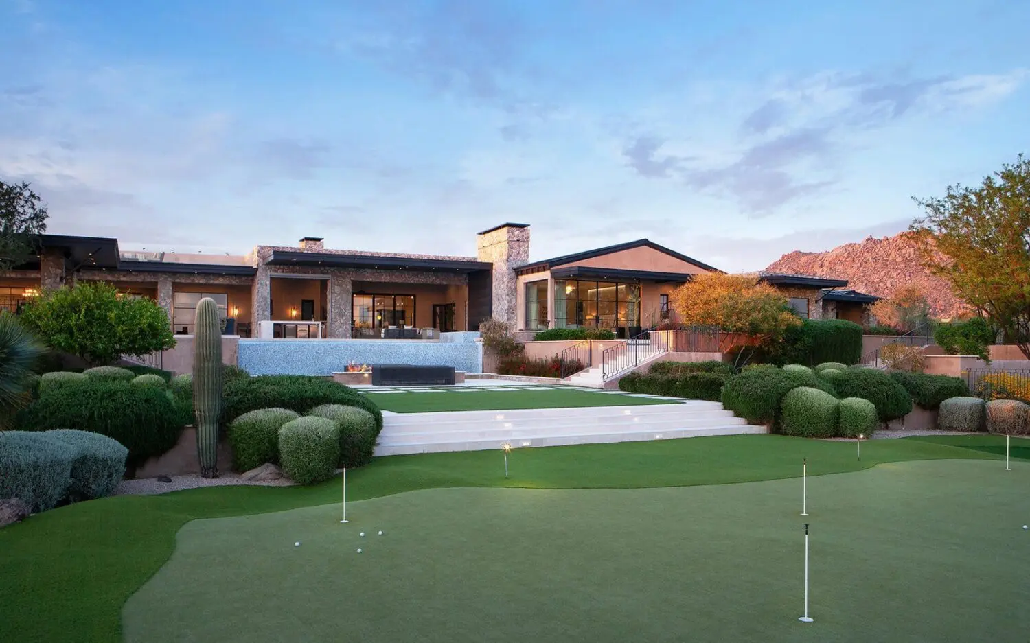 A luxurious estate with a modern design features a manicured lawn and backyard golf greens in the foreground. The house has large windows, a stone chimney, and surrounding shrubbery. The sky is partially cloudy, with mountains in the background.