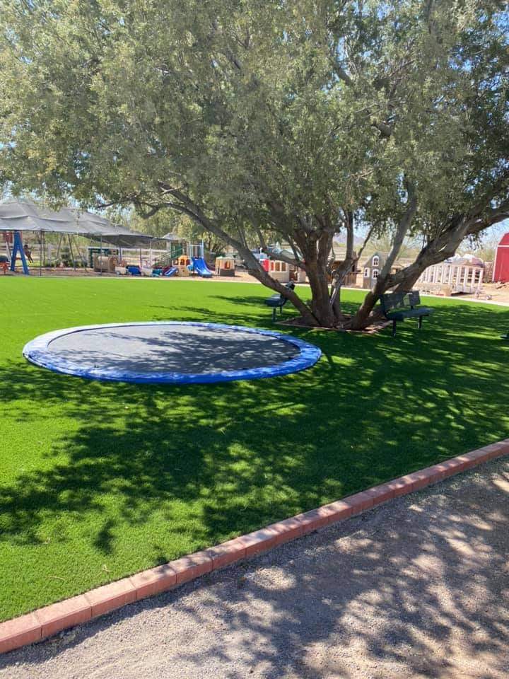 A playground with lush artificial grass is shown, featuring a trampoline set flush with the ground beneath a large tree. Play structures are visible in the background, and two benches are placed nearby. This sustainable area is bordered by a paved path.