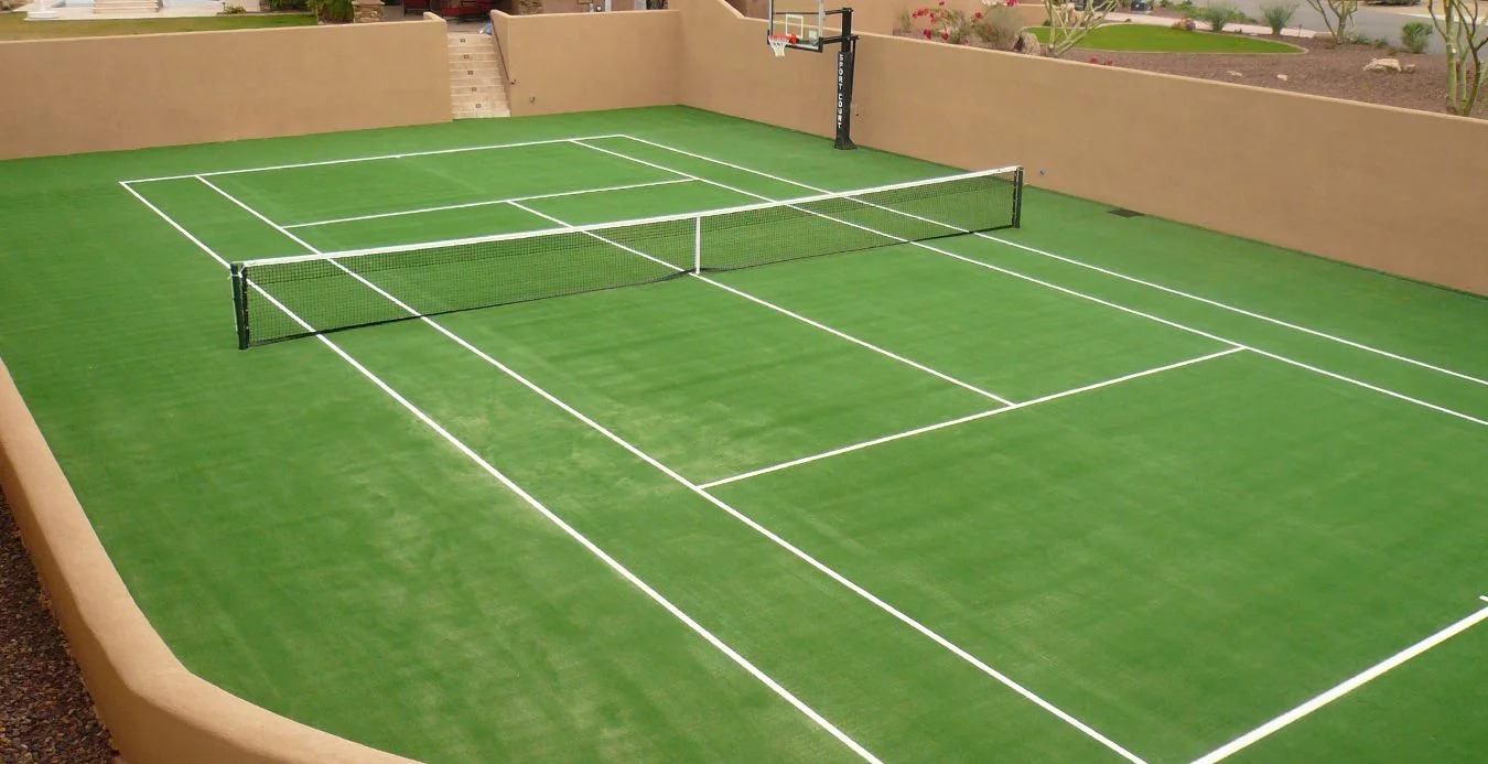 A sunlit pickleball court with white boundary lines and a net positioned in the center. The court, surrounded by beige walls, features sparse desert landscaping and Tucson's signature synthetic grass, while a basketball hoop is visible in the background.