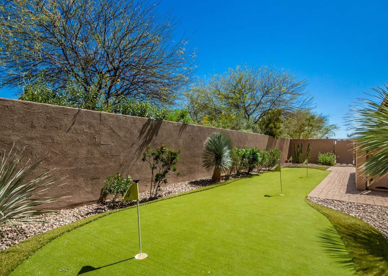 A lush backyard golf green with three flags, featuring expert-installed artificial turf surrounded by desert landscaping including cacti and drought-resistant plants. The area is enclosed by a tall stucco wall, with a clear blue sky overhead providing a sunny backdrop.