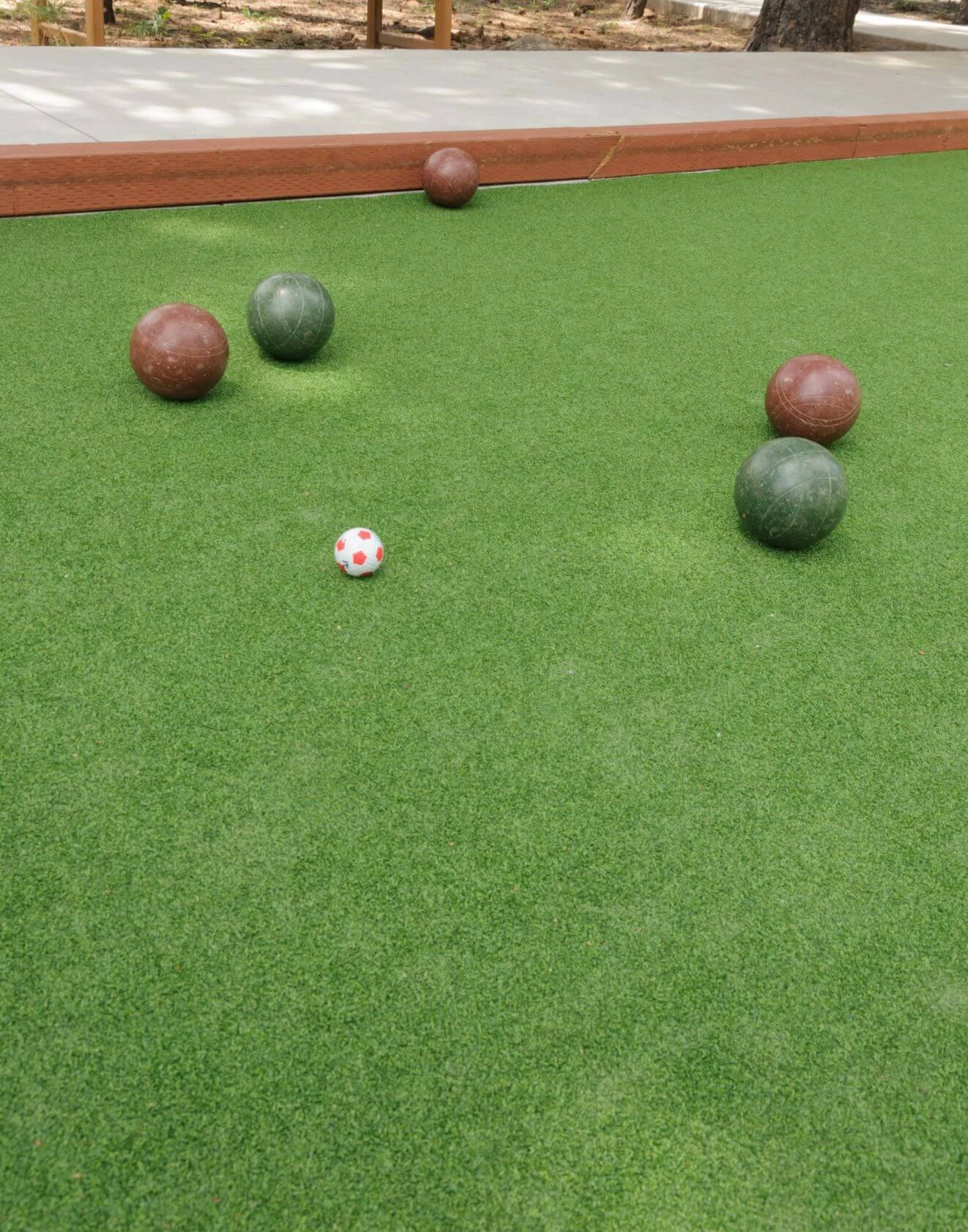 A bocce ball court with several bocce balls scattered on premium artificial turf. The brown and green balls surround a small red and white target ball, meticulously positioned near the wooden boundary. Expertly crafted artificial turf installations offer a perfect playing surface.