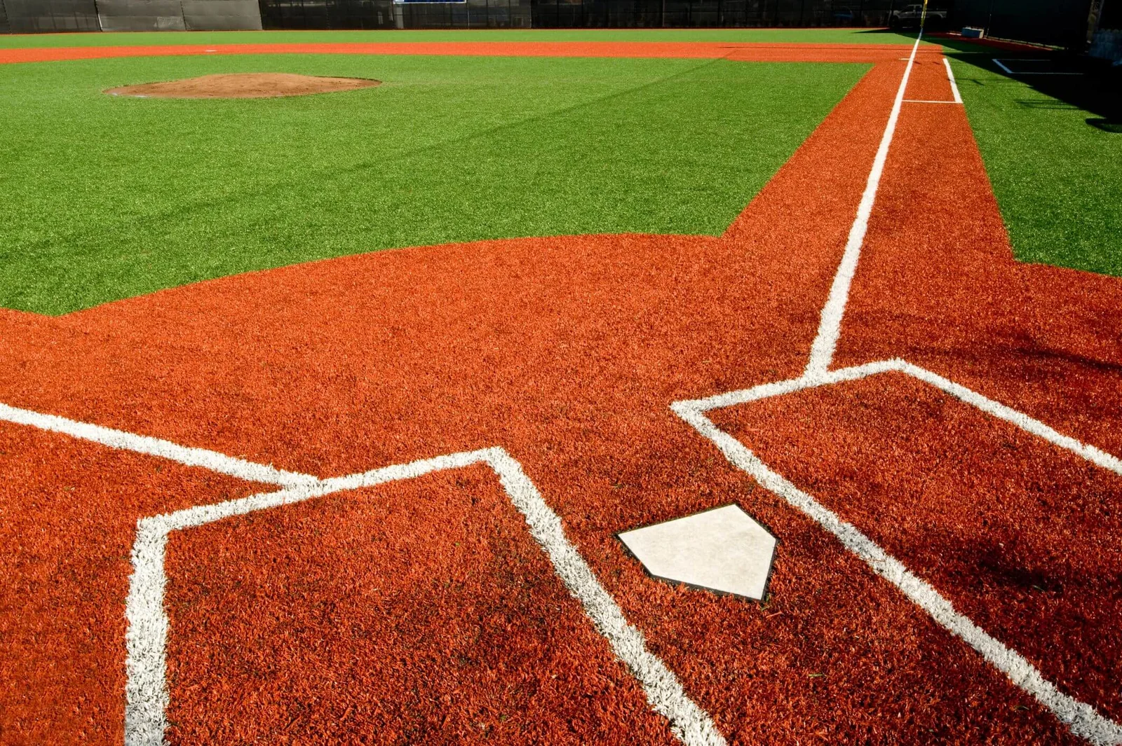 A baseball field in Tucson showcases vibrant green and orange synthetic grass solutions. The image focuses on home plate and the surrounding batter's box, with white lines marking areas on the field. The pitcher's mound stands prominently in the background, highlighting expert artificial turf installations.