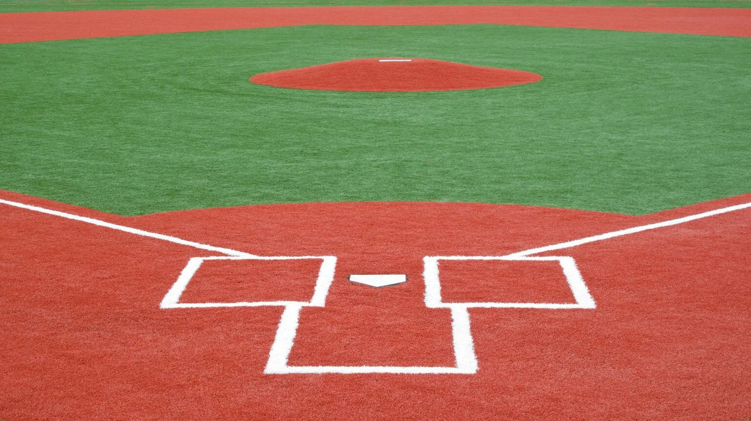 Image of a baseball field's home plate area with a red infield and green outfield made using synthetic grass solutions. The white lines outline the batter's box, foul lines, and pitcher's mound in the distance. Located in Tucson, the field is empty yet ready for action.